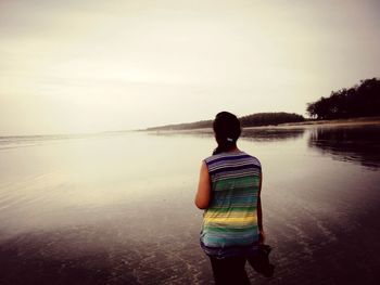 Rear view of woman standing on shore at beach against sky during sunset