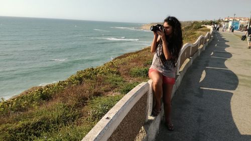 Woman photographing through camera while sitting on retaining wall against sea