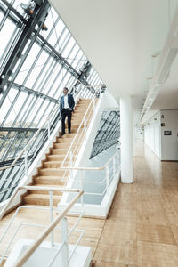 Businessman looking through window while moving down from staircase in office corridor