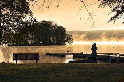 Man sitting on bench by lake against sky during sunset
