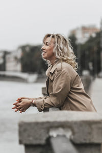 A middle-aged blonde woman with curly hair stands on the river embankment in the city.