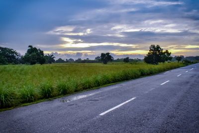 Empty road by trees against sky during sunset