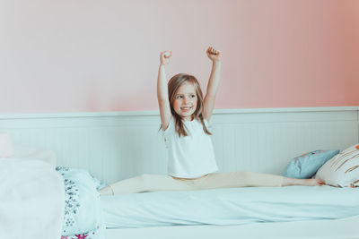 Cheerful girl with arms raised sitting on bed at home