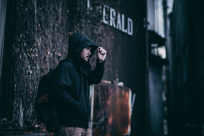 Man looking away while wearing hooded jacket while standing against wall