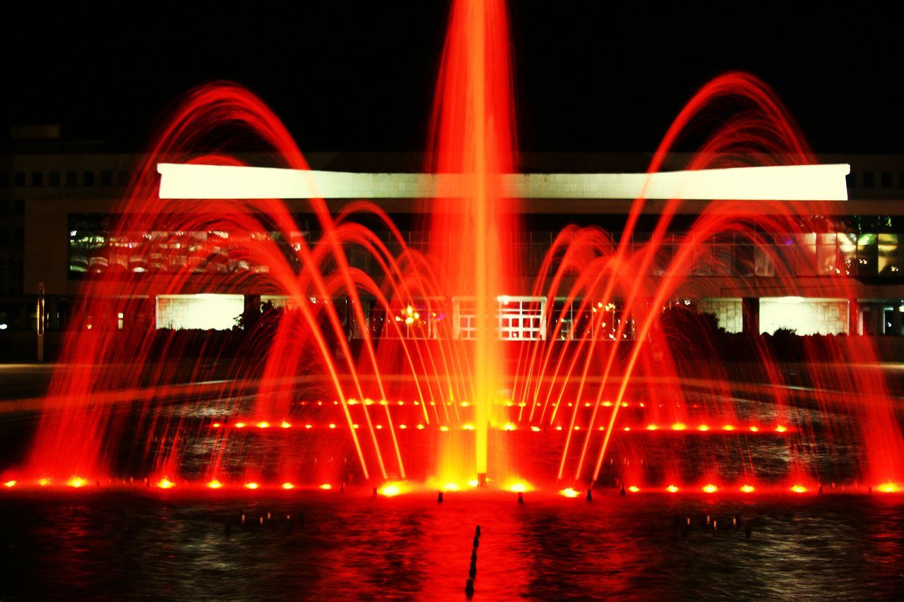 water, reflection, night, illuminated, waterfront, built structure, architecture, building exterior, long exposure, motion, red, river, fountain, arts culture and entertainment, arch, outdoors, blurred motion, multi colored, city, glowing