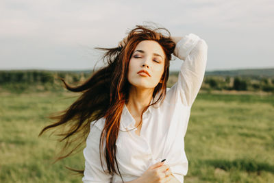 Beautiful young woman with hand in hair standing on land against sky