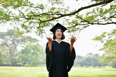 Happy young woman in graduation gown standing on field