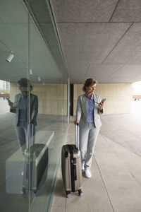 Businesswoman with suitcase using mobile phone standing by glass wall