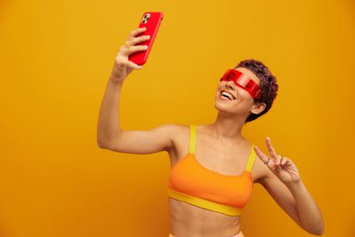 Portrait of smiling young woman taking selfie against yellow background