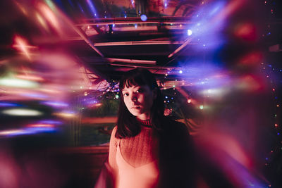 Portrait of young woman standing amidst illuminated multi colored lights at bar