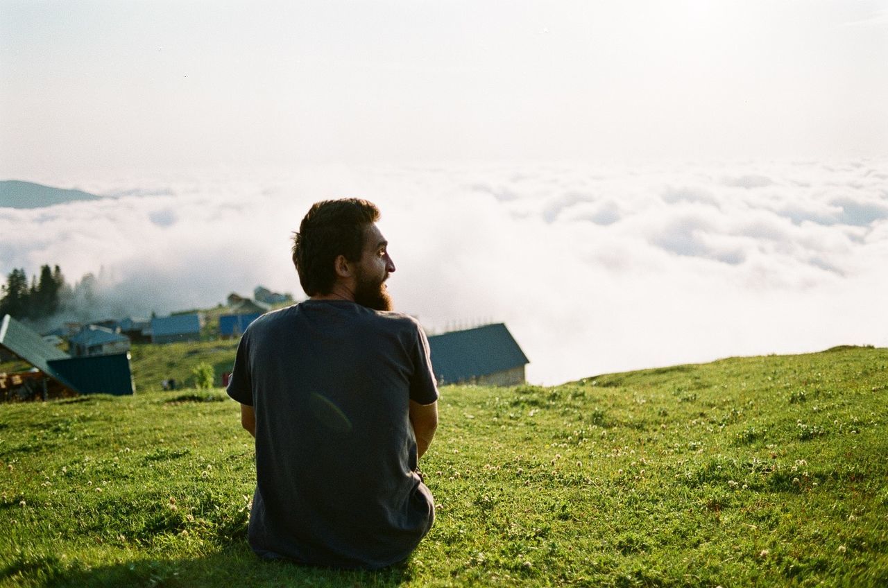 one person, sky, grass, nature, adult, hill, landscape, environment, plant, cloud, men, land, leisure activity, morning, contemplation, beauty in nature, field, rural scene, day, rural area, tranquility, relaxation, scenics - nature, looking, rear view, casual clothing, outdoors, young adult, sunlight, vacation, lifestyles, waist up, trip, green, copy space, plain, holiday, clothing, person, sitting, idyllic, tree, architecture, solitude, technology, meadow, travel, tranquil scene, activity