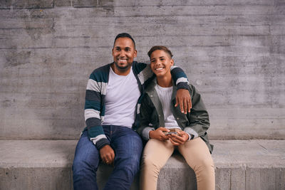 Cheerful father and son looking away while sitting against wall
