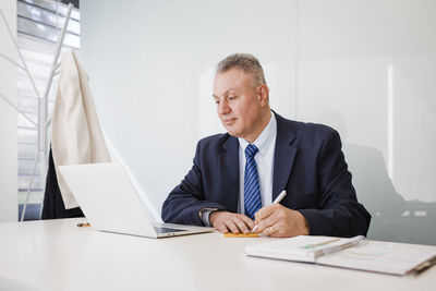 Mature businessman using laptop writing in sticky note at desk in office