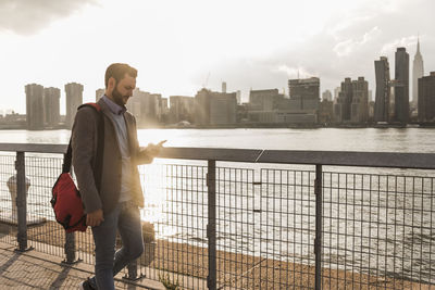 Usa, new york city, businessman walking along east river looking at cell phone