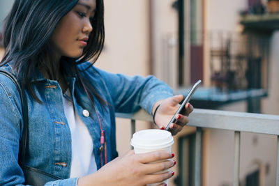 Teenage girl using smart phone while holding disposable coffee cup by railing