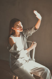 Young woman shielding eyes while sitting on chair against wall