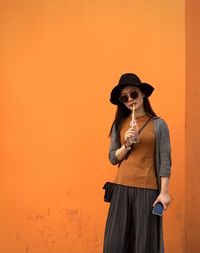 Young woman wearing sunglasses standing against orange wall