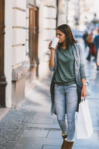 Full length of young woman having coffee while walking on sidewalk in city