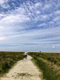 Rear view of man walking on dirt road against cloudy sky