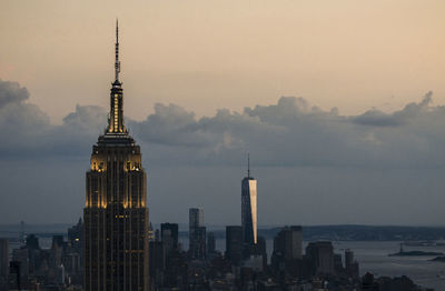 Illuminated empire state building by one world trade center in city against sky during sunset