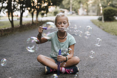 Girl with cross-legged blowing bubbles while sitting on road
