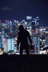 Rear view of silhouette man standing on building terrace against illuminated cityscape at night