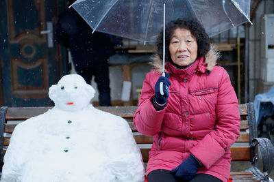 Portrait of woman holding umbrella while sitting snowman on bench