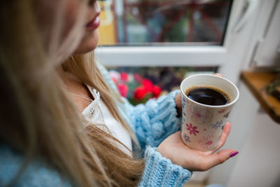 A woman sits at home holding a mug of hot coffee in her hands.
