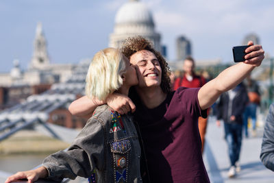 Smiling man with girlfriend taking selfie with mobile phone on footbridge in city