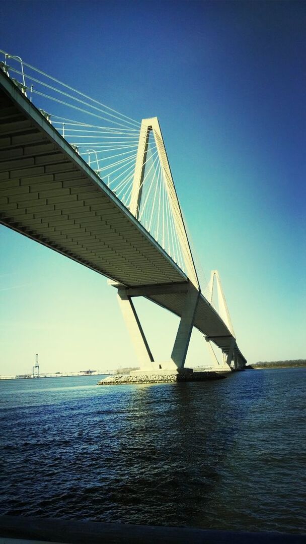 clear sky, water, sea, built structure, connection, blue, transportation, architecture, engineering, suspension bridge, bridge - man made structure, waterfront, bridge, copy space, travel destinations, low angle view, sunlight, outdoors, ocean, travel