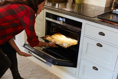 Side view of mature woman inserting dough in oven at home