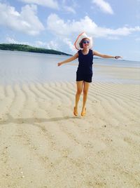 Full length of smiling woman in hat with arms outstretched at beach against sky