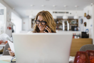 Happy businesswoman with laptop talking on smart phone at restaurant