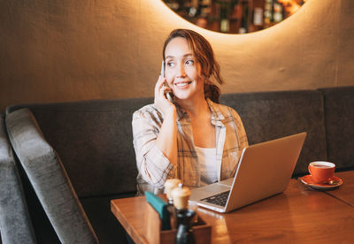 Adult charming brunette woman in plaid shirt working with laptop using mobile phone at cafe