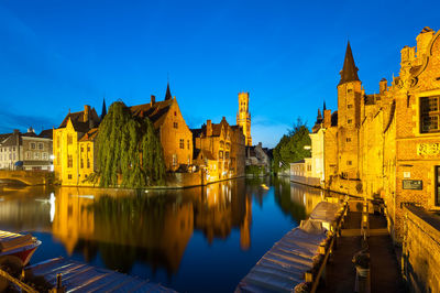 View of illuminated buildings by river against blue sky in city