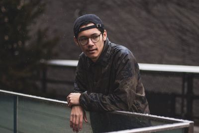 Thoughtful young man looking away while standing by railing during rainfall