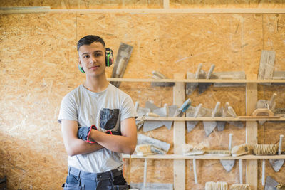 Portrait of confident teenage trainee standing with arms crossed against wall at workshop