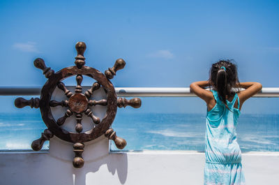 Rear view of girl looking at sea while standing by railing in boat against blue sky