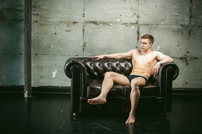 Shirtless muscular man sitting on armchair by wall