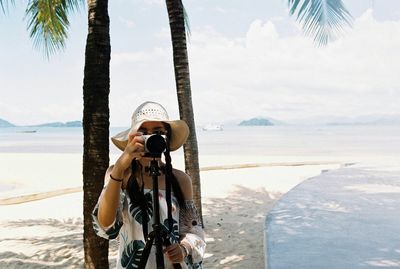 Woman photographing while standing at beach against cloudy sky