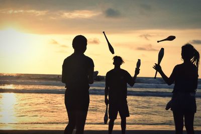 Friends practicing juggling pins at beach against sky during sunset