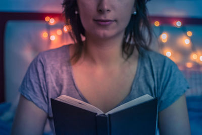 Midsection of young woman reading book while sitting on bed at home