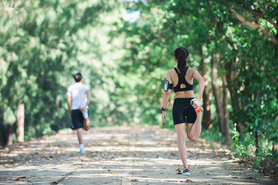 Full length rear view of woman and man stretching leg while exercising on road amidst trees