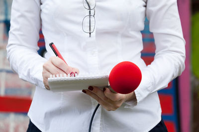 Midsection of female journalist writing on spiral notebook while holding microphone