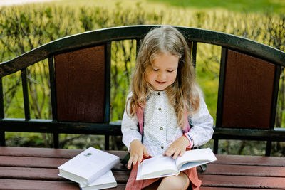 A girl with a backpack is sitting on a bench and reading a book near the school. back to school