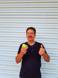 Portrait of smiling mature man having apple while standing against shutter