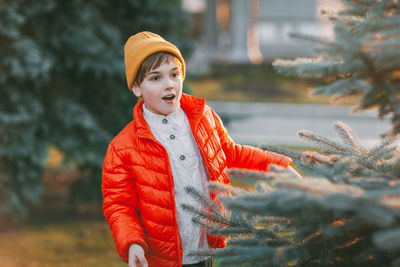 A charming boy walks in the park, happily looks out from behind the christmas tree