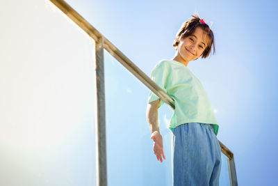 Low angle portrait of girl standing by railing against sky