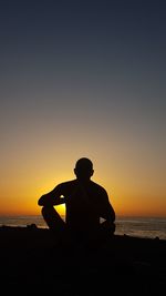 Silhouette man sitting on beach against clear sky during sunset