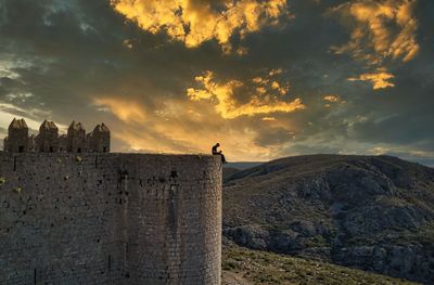 Man sitting on fort against sky during sunset